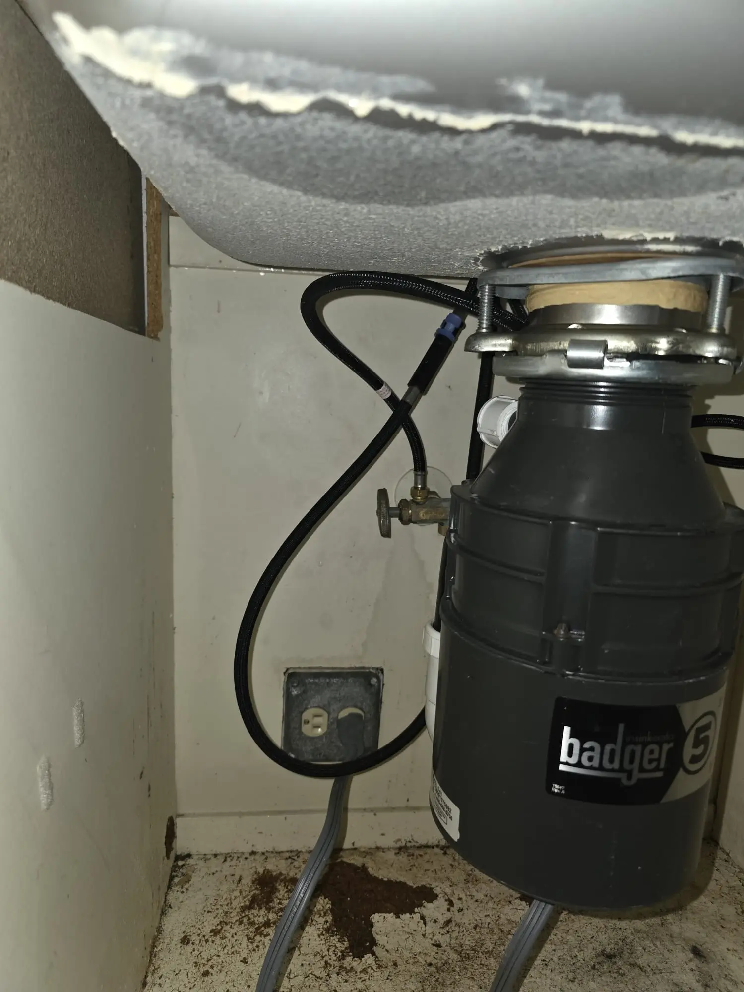 Garbage disposal mounted under a kitchen sink, with a black braided hose, a small shutoff valve and a power cord, and a brown stain on the cabinet floor
