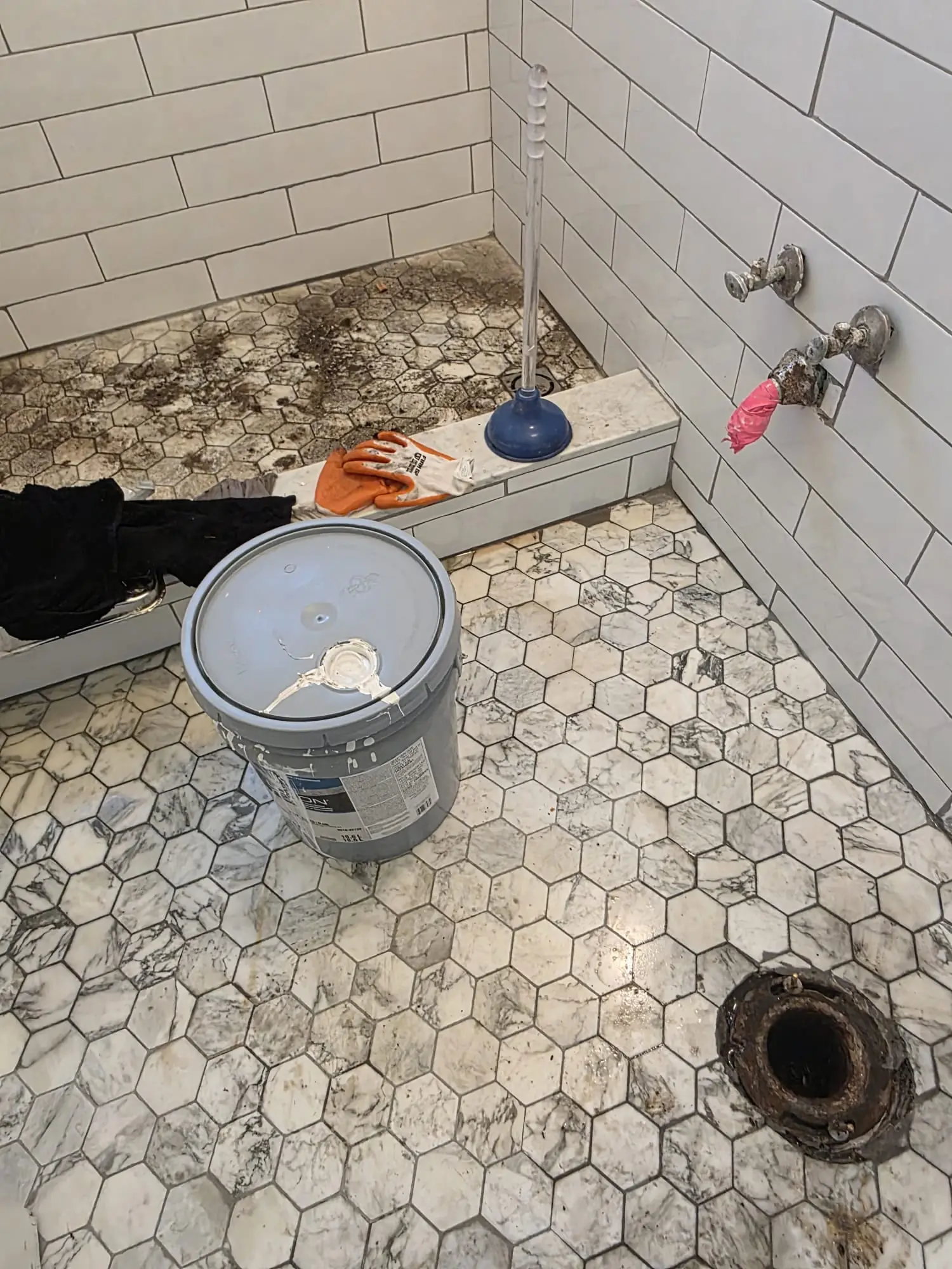 Tiled shower floor with debris around the drain, a plunger and a pair of work gloves on the ledge, a paint bucket on the bathroom floor and an open pipe flange in the floor