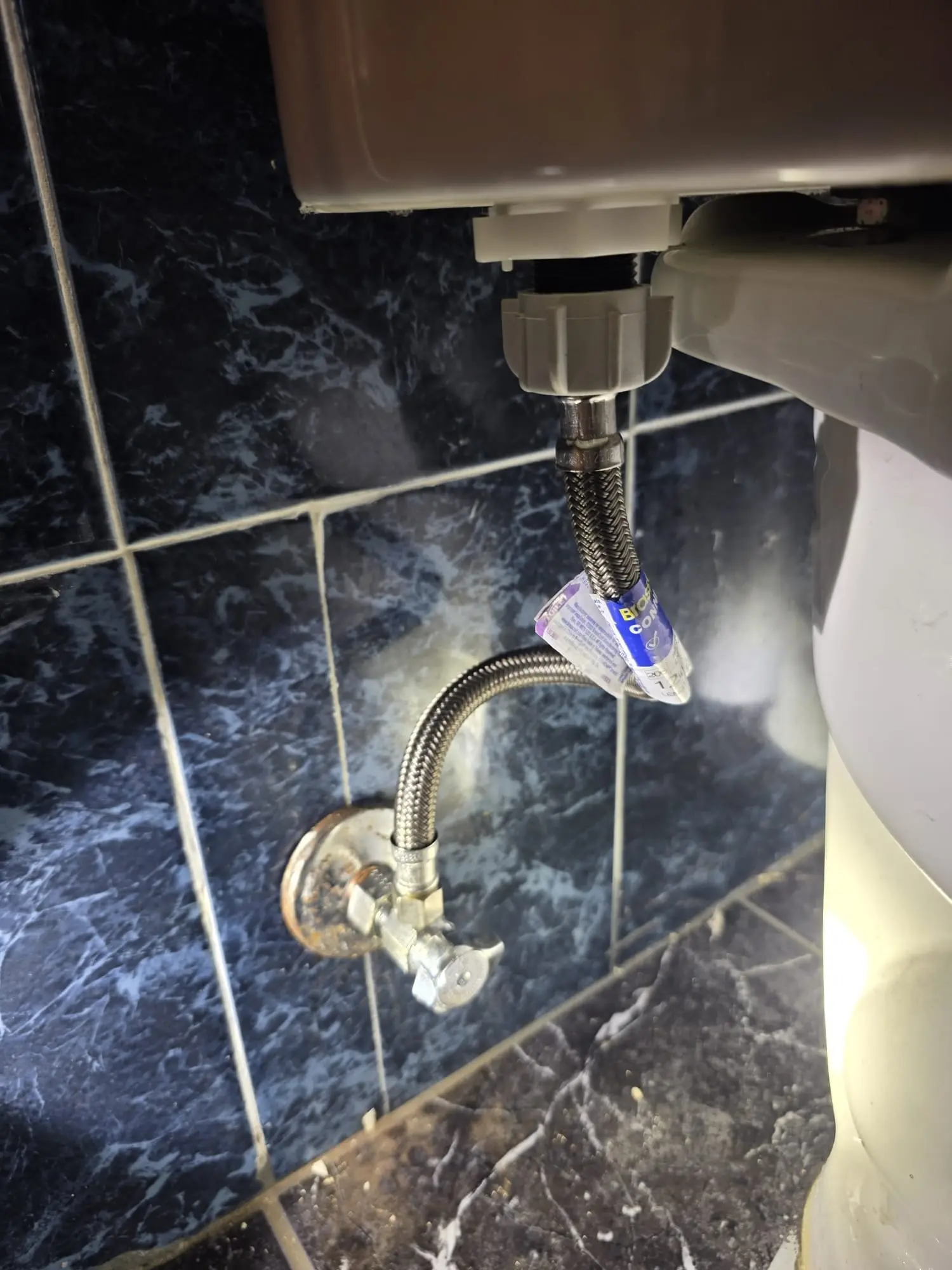 Braided stainless steel supply line running from a corroded angle stop valve on a dark tiled wall up to the underside of a toilet tank
