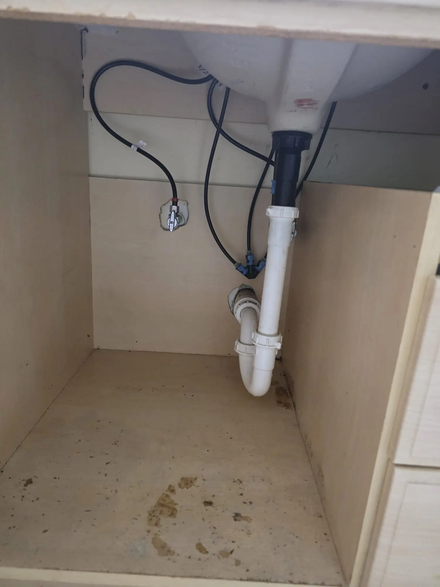 Empty sink cabinet with a white drain trap, two supply lines with small shutoff valves on the back wall and brown stains on the cabinet floor